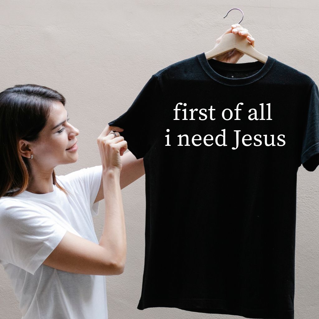 a woman holding a t-shirt with text on that seys "first of all i need jesus"